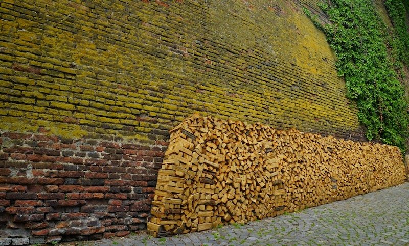 Neatly stacked firewood piled along a weathered moss-covered brick wall with creeping ivy in a quiet cobblestone alley in Prague, Czech Republic