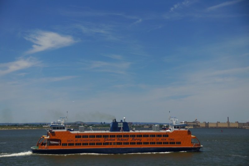 The bright orange Staten Island Ferry glides across New York Harbor on a clear summer day, offering scenic views of the Manhattan skyline and Statue of Liberty. This iconic free ferry connects Lower Manhattan with Staten Island and is a must-do NYC experience