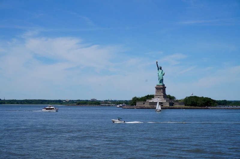 Panoramic view of the Statue of Liberty (La Liberté éclairant le monde) on Liberty Island in New York Harbor, with boats and a sailboat passing in the foreground on a clear summer day. This iconic NYC landmark offers one of the best city vantage points