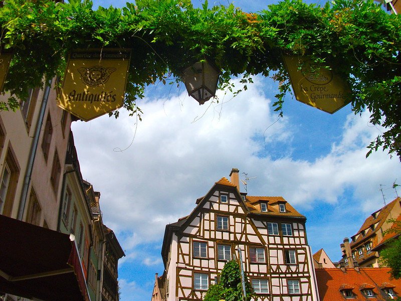 View of Strasbourg’s Old Town in France with half-timbered houses, red tiled rooftops, and leafy greenery overhead framing antique shop and crêperie signs, capturing the charm and mix of French and German influences in the historic district