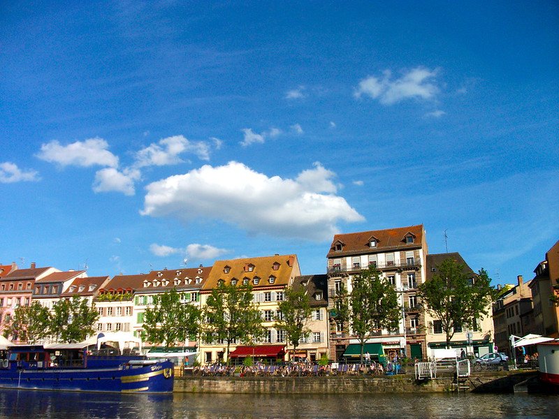 View from a boat tour on the Rhine in Strasbourg, France, with colorful historic houses, riverside cafés, and leafy trees along the waterfront under a bright blue sky, showcasing the city’s unique blend of French and German influences