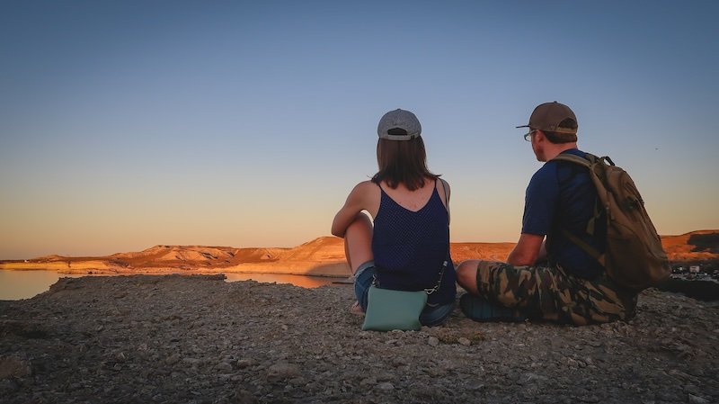 Audrey Bergner of That Backpacker and Nomadic Samuel sit side by side at sunrise, enjoying panoramic views over the cliffs and coastline of Puerto Pirámides, Península Valdés, Argentina, after an early morning hike to a scenic high vantage point