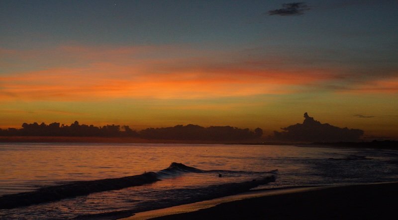 Fiery orange and soft pink hues streak across the horizon as the sun sets over the calm Caribbean Sea, with gentle waves lapping the darkened shoreline in Cayo Santa María, Cuba, creating a romantic and serene honeymoon evening scene