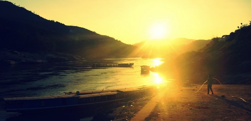 Golden sunset over the Mekong River in Laos with long, narrow slow boats moored along the shore and silhouettes of distant mountains on the horizon. The warm light bathes the riverbank, capturing the tranquil atmosphere at the end of a day’s journey toward Luang Prabang
