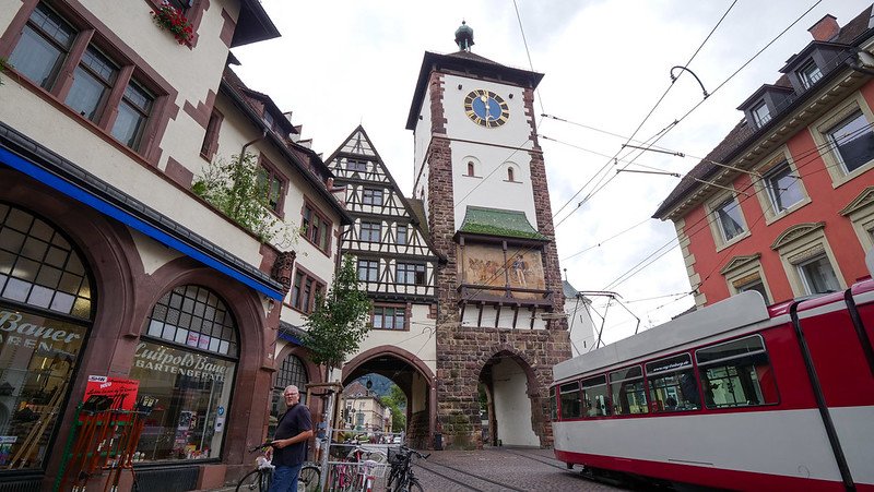 Historic Swabian Gate (Schwabentor) in Freiburg im Breisgau, Germany, featuring its medieval clock tower, half-timbered buildings, and a passing red tram along cobblestone streets