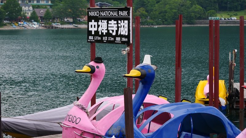Colourful swan-shaped pedal boats docked along the shore of Lake Chuzenji in Nikko National Park, Nikko, Japan, with a sign displaying the lake’s elevation and depth in the background