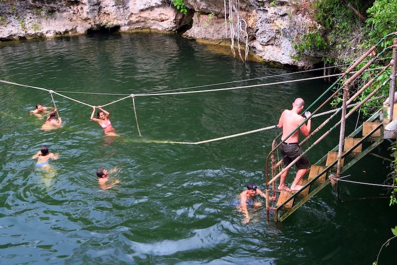 Travelers cooling off while swimming in a natural cenote near Playa del Carmen, surrounded by rocky cliffs and jungle vegetation, with some climbing a ladder and others holding onto ropes stretched across the water Travelers cooling off while swimming in a natural cenote near Playa del Carmen, surrounded by rocky cliffs and jungle vegetation, with some climbing a ladder and others holding onto ropes stretched across the water