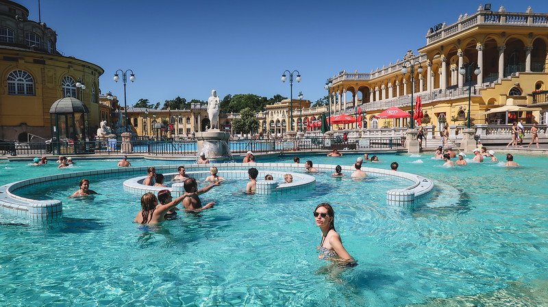 Audrey Bergner of That Backpacker enjoying a sunny day at the iconic Széchenyi Thermal Baths in Budapest, Hungary. Surrounded by historic yellow buildings and other visitors, she relaxes in the steaming outdoor pool—one of the city’s top attractions