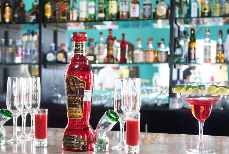 Vibrant tequila tasting setup at a Playa del Carmen bar, featuring a striking red bottle of Tequila Castellano, lime-garnished cocktails, and shot glasses against a colorful backdrop of assorted spirits