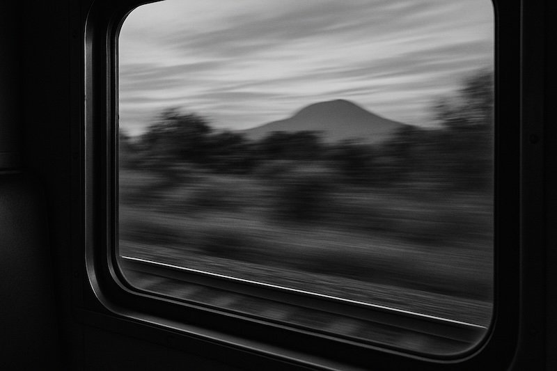 A black and white photograph taken from inside a moving Thai train, looking out the window at a blurred rural landscape with trees, fields, and a distant mountain. The sharp window frame contrasts with the motion-blurred scenery, creating a nostalgic travel atmosphere