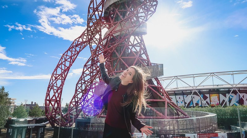 That Backpacker Audrey Bergner poses happily in front of the ArcelorMittal Orbit in London’s Queen Elizabeth Olympic Park, with the sun shining behind the red looping structure that features the world’s longest tunnel slide attraction