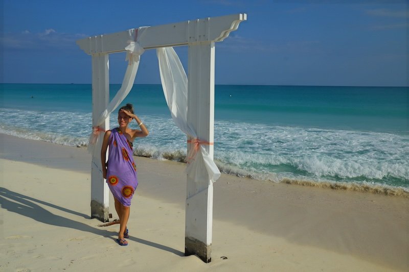 That Backpacker Audrey Bergner stands beneath a flowing white beach arch on the soft sand, gazing out toward the turquoise waves of Cayo Santa María, Cuba, during a serene and sun-soaked honeymoon stroll by the sea.