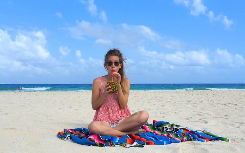 That Backpacker Audrey Bergner enjoying a tropical pineapple drink while relaxing on a colorful Mexican blanket at the beach in Playa del Carmen, with turquoise waves and blue skies in the background