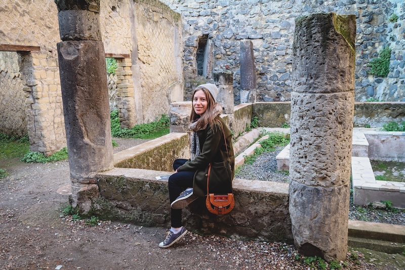 That Backpacker Audrey Bergner bundled up and layers and wearing a beanie taking a break to sit down visiting Herculaneum, Italy 