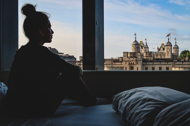 That Backpacker Audrey Bergner sits by the window at CitizenM Hotel in London, gazing out at the historic Tower of London with its medieval turrets and Union Jack flag flying, capturing a perfect travel moment blending modern comfort and iconic heritage