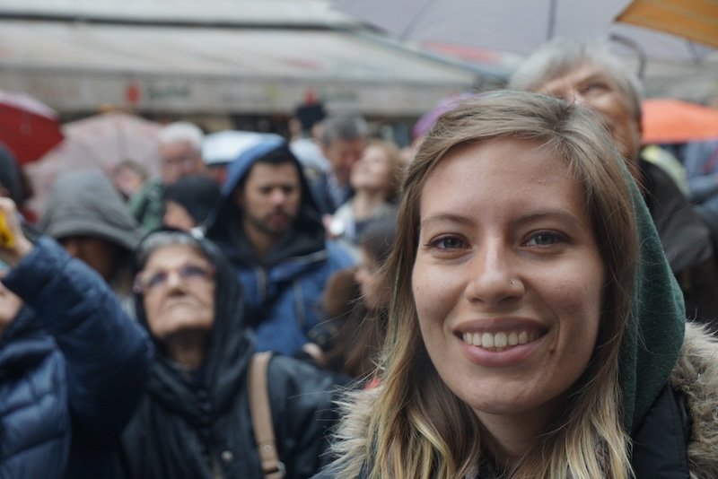 That Backpacker Audrey Bergner smiling in the foreground with a blurred crowd gathered behind her on a rainy day in Prague, Czech Republic, during a popular city event