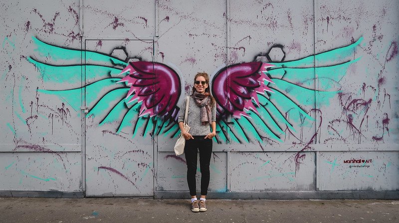 That Backpacker Audrey Bergner poses in front of a vibrant wings mural in East Croydon, London, standing against a grey wall painted with turquoise and magenta feathers, creating a playful street art moment that transforms her into part of the artwork