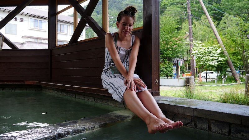 Audrey Bergner of That Backpacker relaxing with her feet soaking in the warm mineral waters of a public foot bath at Yumoto Onsen in Nikko, Japan, surrounded by peaceful mountain scenery