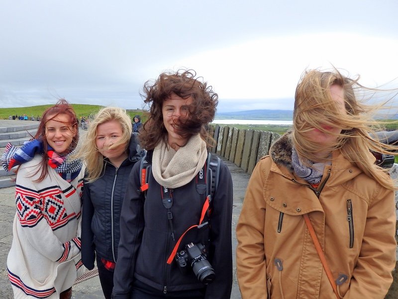 That Backpacker Audrey Bergner standing with her friends and sisters during a windy stop on a Ireland road trip, with everyone’s hair wildly blowing in the gusts as they laugh and pose against a dramatic coastal backdrop, capturing the fun and unpredictable weather of traveling Ireland