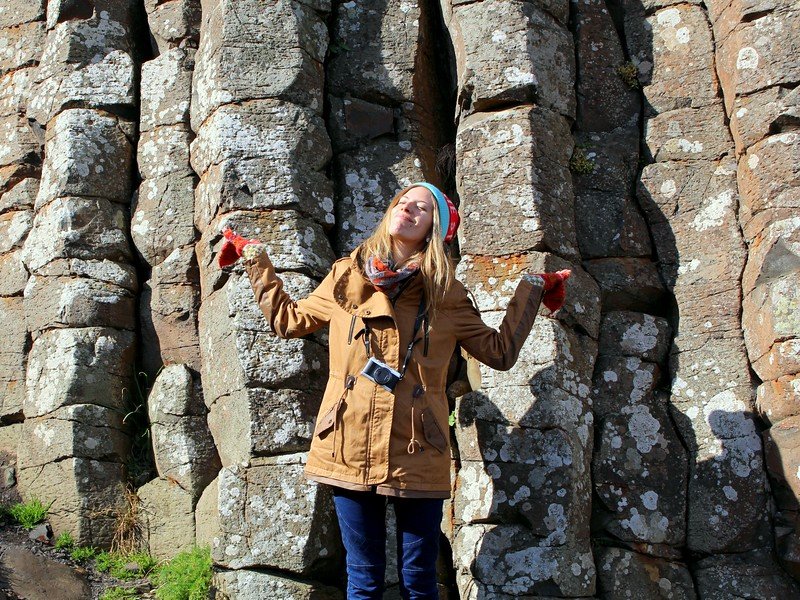 That Backpacker Audrey Bergner smiling with arms raised in joy while standing among the hexagonal basalt columns of Giant's Causeway in Northern Ireland, bundled in a brown coat, scarf, and knit hat during a girls’ road trip adventure along the rugged Atlantic coastline