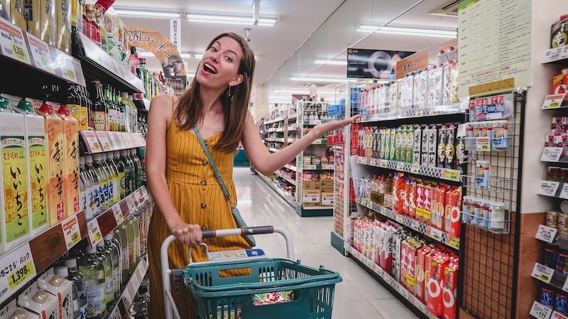 Audrey Bergner of That Backpacker smiling while grocery shopping in Nikko, Japan, pushing a shopping cart down an aisle lined with colourful bottles and cartons of Japanese drinks and condiments