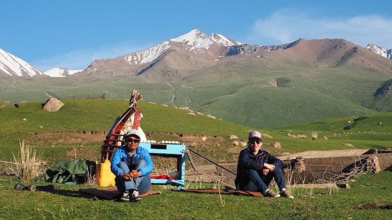 That Backpacker Audrey Bergner sitting on the grass beside our local guide with snow-capped mountains and green pastures in the background during the 3-Day Panorama Trek in Kyrgyzstan