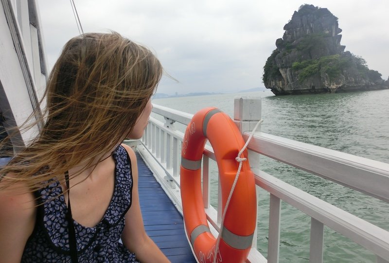 That Backpacker Audrey Bergner sitting on the deck of a wooden tour boat in Halong Bay, Vietnam, with wind blowing through her hair as she gazes toward a towering limestone karst island rising from the calm emerald-green waters, framed by an orange life ring and the boat’s white railings under a cloudy sky