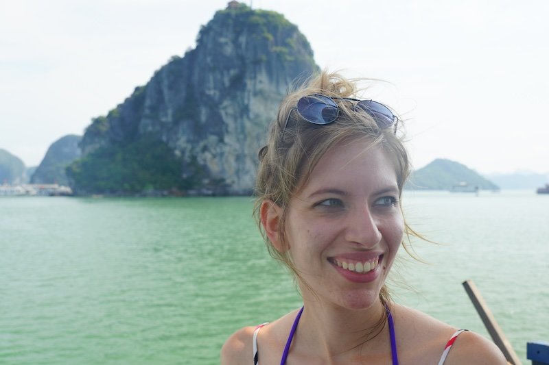 That Backpacker Audrey Bergner smiling aboard a boat in Halong Bay, Vietnam, with towering limestone karst islands rising dramatically from the emerald-green water in the background on a bright and hazy day during a scenic cruise
