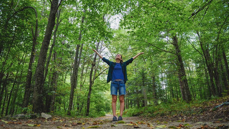 That Backpacker Audrey Bergner standing on a forest trail at Au Diable Vert in Sutton, Quebec, joyfully stretching her arms toward the leafy green canopy while hiking in the Eastern Townships