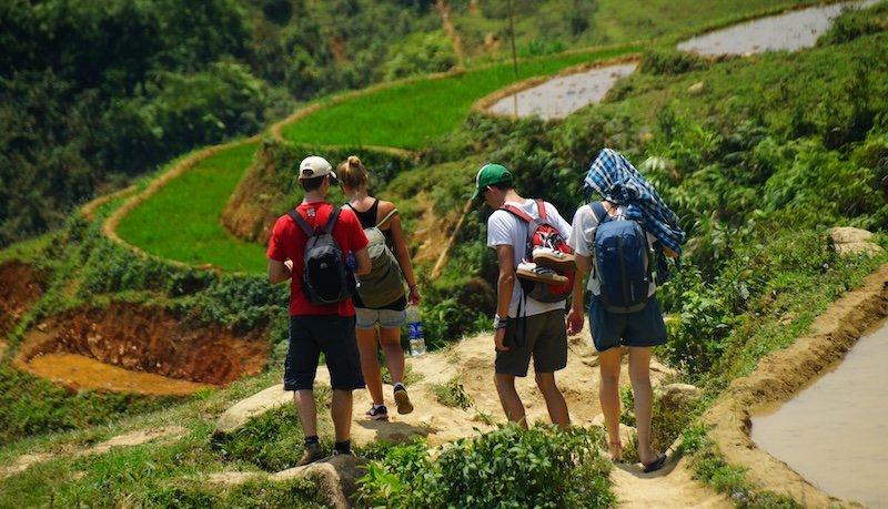 Audrey Bergner from That Backpacker trekking with a small hiking group along narrow dirt paths beside vibrant green rice terraces in Sapa, Vietnam under the midday sun