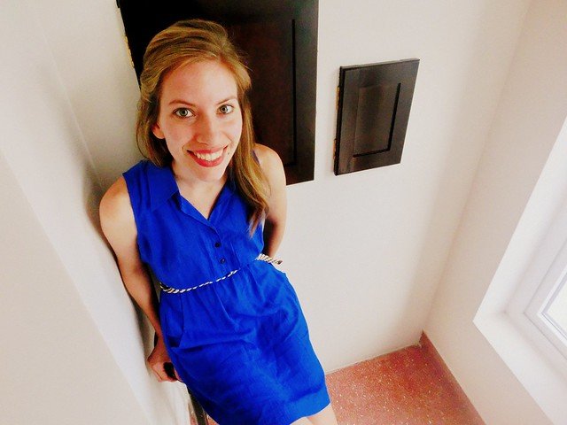 That Backpacker Audrey Bergner smiling while modeling a custom-made royal blue sleeveless dress with a braided belt, tailored by a local Hoi An tailor in Vietnam, standing indoors against a cream-colored wall and showcasing the vibrant bespoke fashion the town is known for