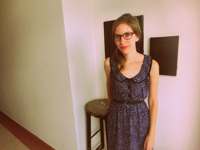 That Backpacker Audrey Bergner wearing a custom-made vintage-inspired navy blue dress with a subtle floral print and a round peter pan neckline, tailored by a local Hoi An tailor in Vietnam, standing indoors against a minimalist cream wall next to a rustic wooden stool