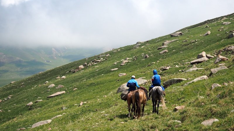 That Backpacker Audrey Bergner riding a horse alongside a local guide through a misty green mountain slope scattered with rocks during the 3-Day Panorama Trek in Kyrgyzstan