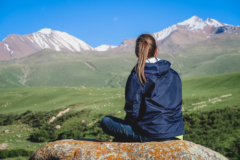That Backpacker Audrey Bergner sitting cross-legged on a rock overlooking the snow-capped peaks and rolling green valleys of the Tian Shan Mountains during the 3-Day Panorama Trek in Kyrgyzstan, taking a break from horse trekking to admire the alpine scenery under a bright blue sky