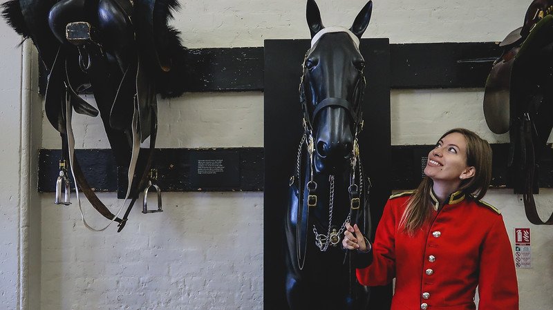 That Backpacker Audrey Bergner smiles while dressed in a red guard’s uniform at the Household Cavalry Museum in London, standing beside a black horse display and saddles, enjoying an interactive experience with Britain’s ceremonial traditions