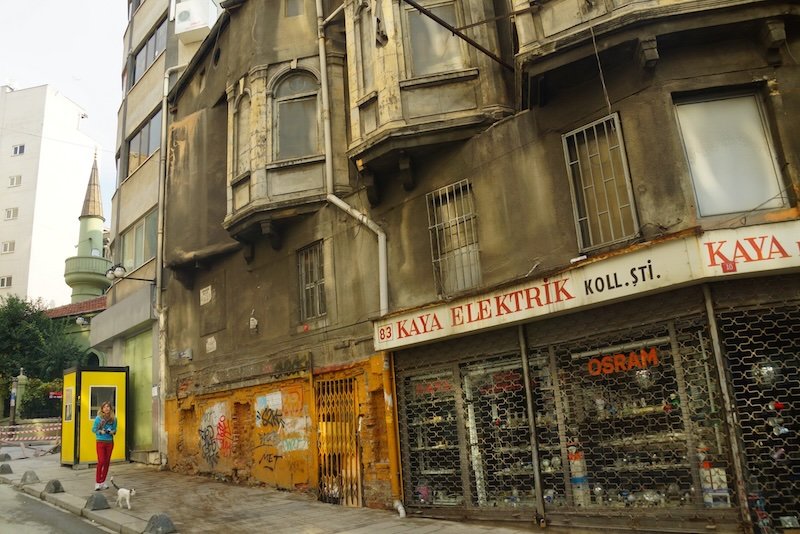That Backpacker Audrey Bergner standing beside a bright yellow phone booth on a quiet side street in Istanbul, Turkey, next to an old weathered building with shuttered shops, colorful graffiti, and a curious stray cat walking nearby