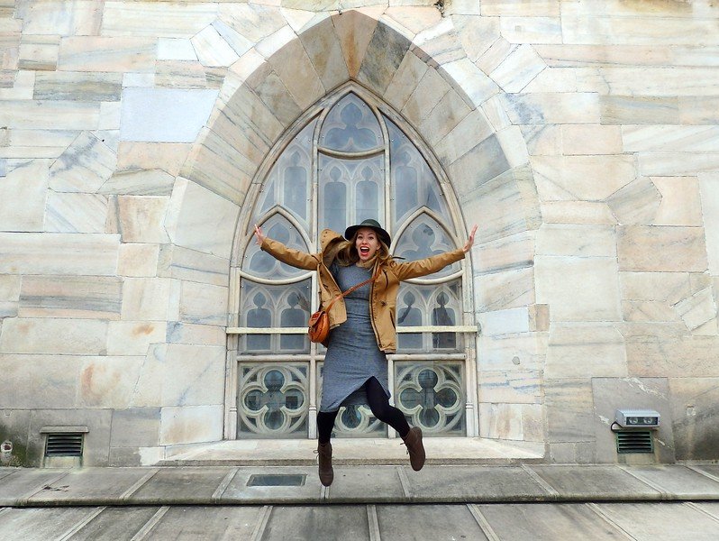 That Backpacker Audrey Bergner jumping with joy in front of a stunning Gothic arched window on the rooftop terraces of Milan Cathedral (Duomo di Milano) during a city break in Milan, Italy