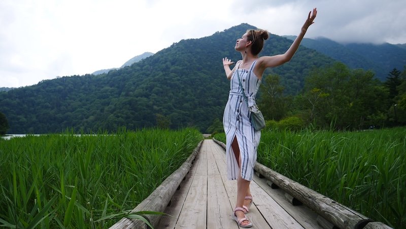Audrey Bergner of That Backpacker joyfully standing on a wooden boardwalk surrounded by lush green marsh grasses near Lake Yunoko in Nikko, Japan, with misty forested mountains in the background