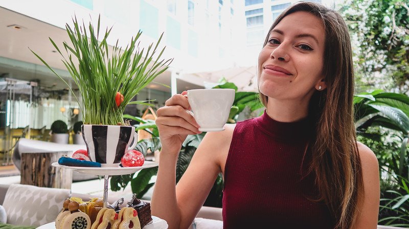 That Backpacker Audrey Bergner enjoys Mad Hatter’s Afternoon Tea in London, smiling while holding a teacup beside a whimsical tiered tray of colorful pastries and treats, capturing the playful Alice in Wonderland-inspired dining experience