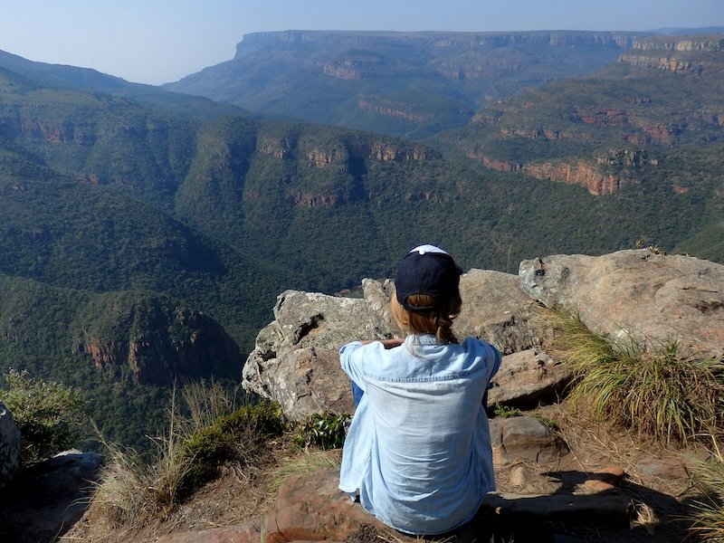 That Backpacker Audrey Bergner sitting on a rocky cliff edge wearing a cap and light long-sleeve shirt while admiring the sweeping green mountain vistas of South Africa during a safari adventure