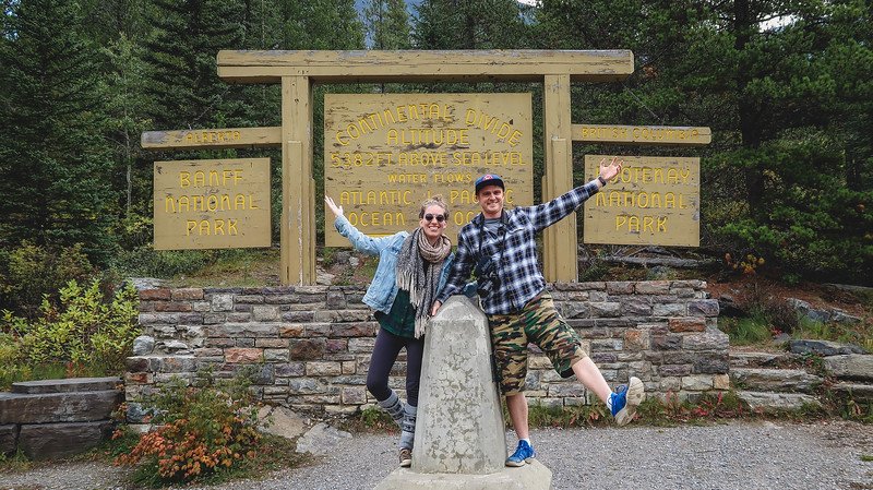Audrey Bergner from That Backpacker and Nomadic Samuel posing joyfully at the Continental Divide marker between Banff National Park and Kootenay National Park in the Rocky Mountains, where water flows toward both the Atlantic and Pacific Oceans