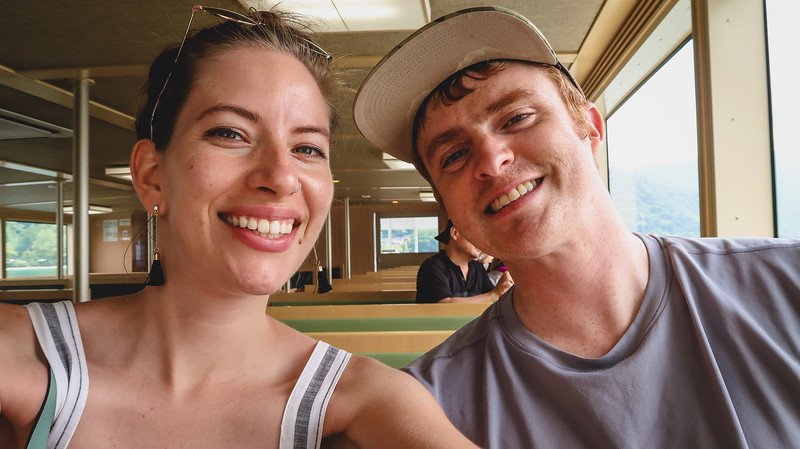 Nomadic Samuel and Audrey Bergner of That Backpacker smiling during a scenic boat tour on Lake Chuzenji in Nikko, Japan, sitting inside the ferry with lake views through the windows behind them