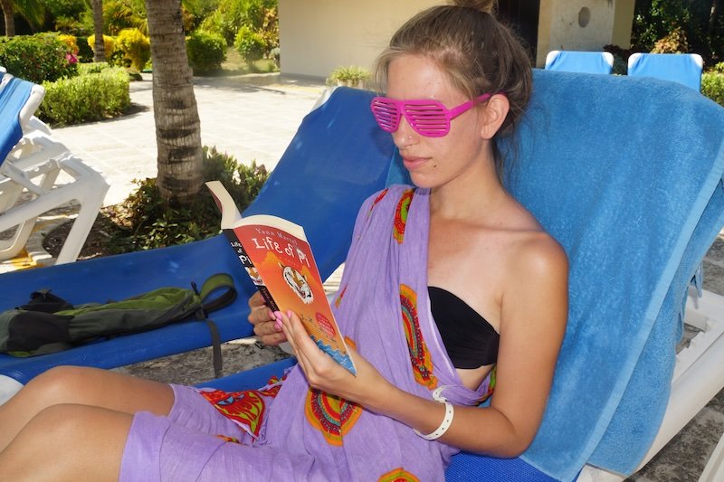 That Backpacker Audrey Bergner lounges poolside on a blue sun chair wearing pink shutter shades and a purple sarong while reading Life of Pi by Yann Martel during a relaxing honeymoon afternoon at a resort in Cayo Santa María, Cuba