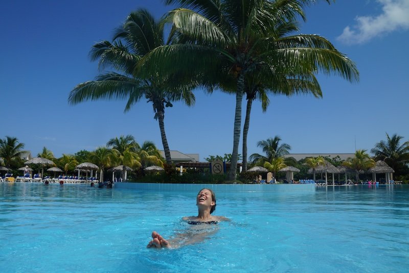 Audrey Bergner from That Backpacker floats joyfully in a turquoise resort pool surrounded by tall palm trees and palapa umbrellas under a sunny blue sky in Cayo Santa María, Cuba during a relaxing honeymoon getaway