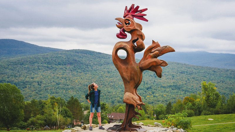 That Backpacker Audrey Bergner admiring a giant whimsical rooster sculpture with panoramic views of the Green Mountains of Vermont in the distance from Au Diable Vert in Sutton, Quebec, a scenic spot in the Eastern Townships