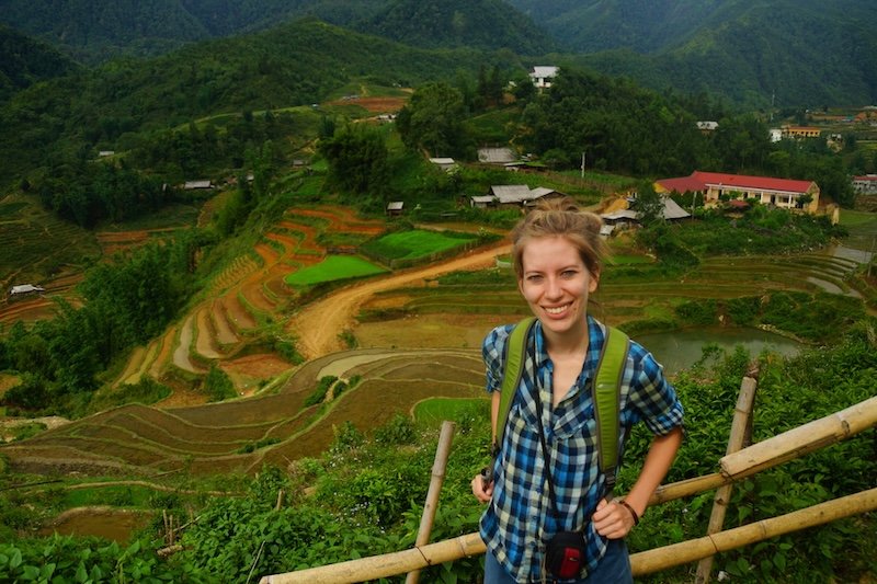 That Backpacker Audrey Bergner smiling during a trek in the terraced hills of Sapa, Vietnam, wearing layered hiking clothes and carrying a green backpack as she stands on a bamboo-lined trail overlooking colorful rice paddies, scattered hilltop houses, and lush green mountains under a cloudy sky