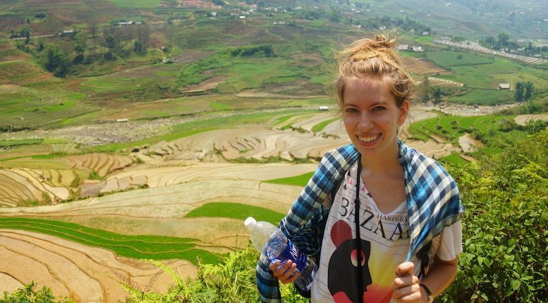 Audrey Bergner from That Backpacker smiling while holding a water bottle during a trek in Sapa, Vietnam, standing above terraced rice fields and wearing layered clothing for the changing mountain weather