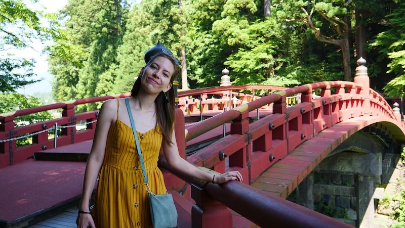 Audrey Bergner of That Backpacker smiling in a mustard-yellow dress while standing on the iconic vermilion Shinkyo Bridge in Nikko, Japan, surrounded by lush green forest