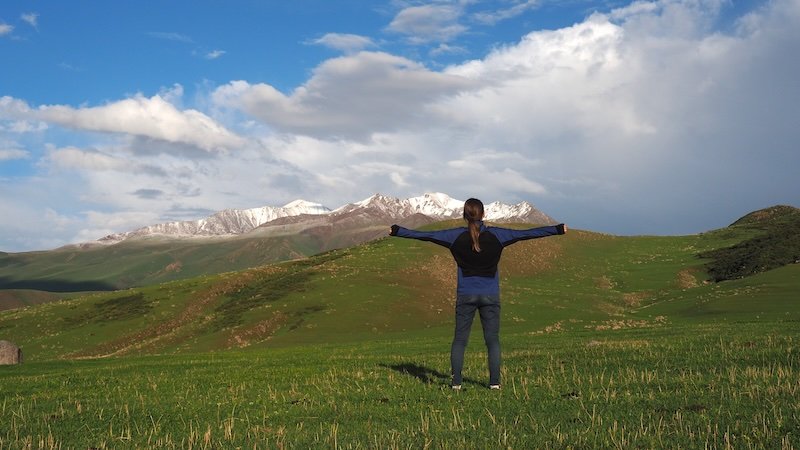 That Backpacker Audrey Bergner standing with arms outstretched on a grassy hillside, gazing at the snow-capped Tian Shan Mountains during the 3-Day Panorama Trek in Kyrgyzstan