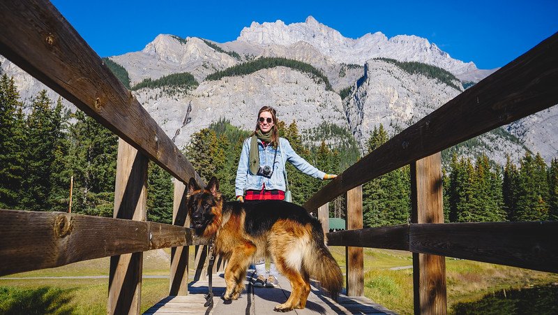 Audrey Bergner from That Backpacker standing on a wooden bridge with Togo the German Shepherd at Cascade Ponds, surrounded by evergreen forest and towering rocky peaks in the Canadian Rockies on a sunny day.